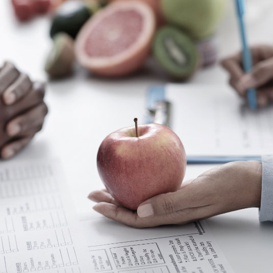 Woman's hand holding apple with forms from a medical weight loss program on the table